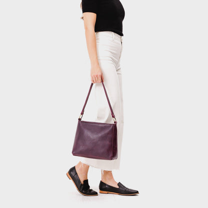 Plum*Classic | Model holding leather triangular shoulder bag with zipper top closure and wide shoulder strap